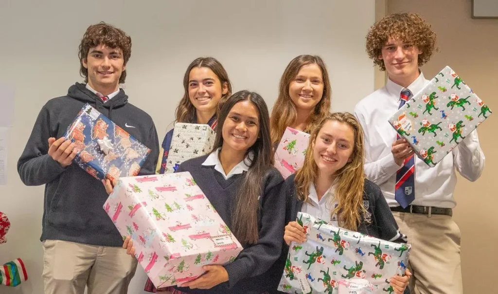 The image depicts a group of six high school students, smiling and holding colorful wrapped gifts with festive holiday designs. The group appears cheerful and is likely participating in a gift-giving or holiday event. They are dressed in school uniforms or casual attire.