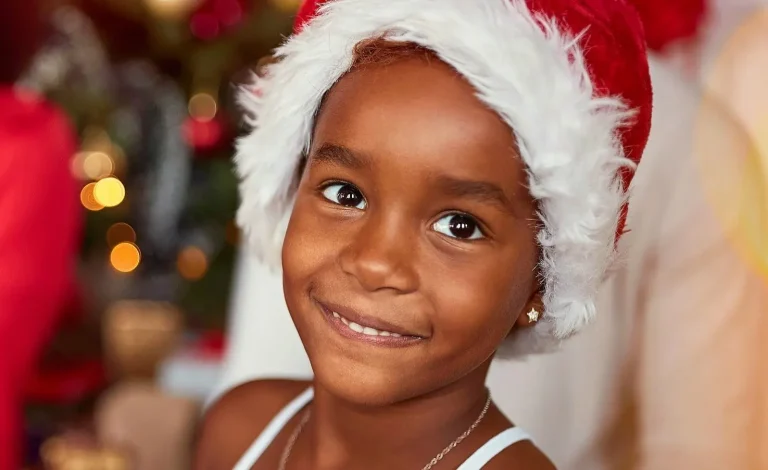 The image shows a smiling child wearing a festive Santa hat, radiating joy and warmth. The blurred background features colorful holiday lights, evoking a cheerful and celebratory atmosphere, possibly related to a seasonal or charitable event.